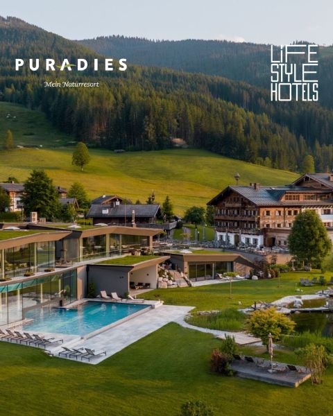 Modernes Hotel mit Außenpool neben traditionellen Alpenhäusern, gelegen in einem üppig grünen Tal, umgeben von bewaldeten Hügeln.