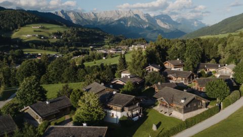 Luftaufnahme eines Dorfes mit Holzhäusern, umgeben von grünen Hügeln, Bäumen und Bergen unter einem teilweise bewölkten Himmel.
