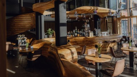 Modernes Bar-Interieur mit geschwungenen Holztheken, Hängeleuchten, ausgestellten Flaschen, runden Tischen und braunen Stühlen im natürlichen Sonnenlicht.