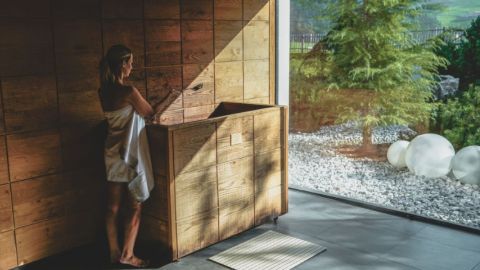 Eine Frau in einem Handtuch steht neben einer hölzernen Badewanne in einem modernen, sonnendurchfluteten Wellnessraum mit einem großen Fenster, das den Blick auf einen Garten freigibt.