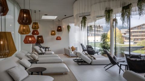 Moderne Lounge mit weißen Liegesesseln, geometrischen Hängelampen, raumhohen Fenstern und Pflanzen, mit malerischem Blick ins Freie.