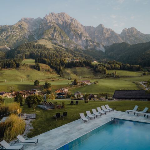 Ein Swimmingpool mit Liegestühlen bietet einen Blick auf ein grünes Tal, vereinzelte Häuser und hohe Berge unter einem klaren Himmel.