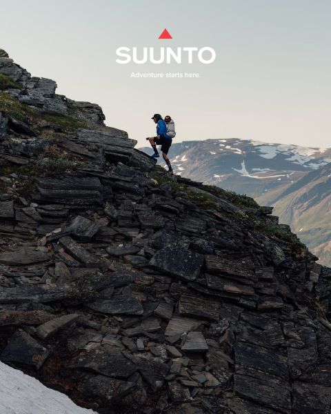 Ein Wanderer erklimmt einen felsigen Berghang mit Schneeflecken, über dem die Aufschrift „SUUNTO Adventure starts here.“ zu lesen ist. Im Hintergrund sind schneebedeckte Gipfel zu sehen.
