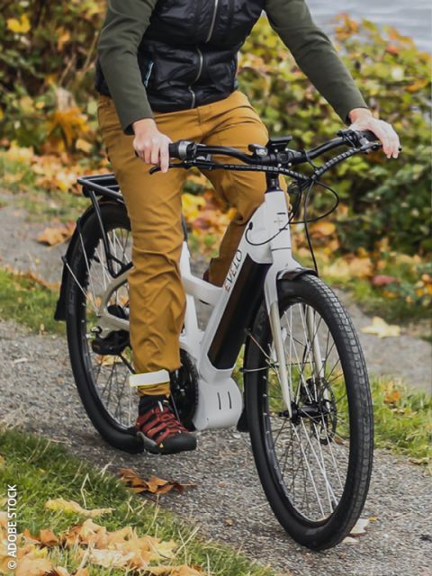 Eine Nahaufnahme eines schwarzen Elektrofahrrads, das auf einer Straße in der Stadt geparkt ist, mit unscharfen Gebäuden und anderen Fahrrädern im Hintergrund, unter einem bedeckten Himmel.