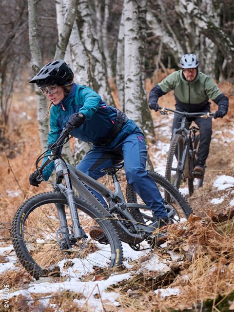 Zwei Radfahrer in Winterkleidung fahren auf einem schmalen, schneebedeckten Weg durch einen Wald mit schlanken Bäumen, durch deren Äste das Sonnenlicht fällt.
