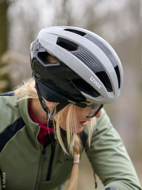 Ein Mann in grüner Jacke und Handschuhen prüft einen schwarzen Fahrradhelm und sitzt vor einer Holzwand. Er trägt ein schwarzes Stirnband mit weißer Schrift.