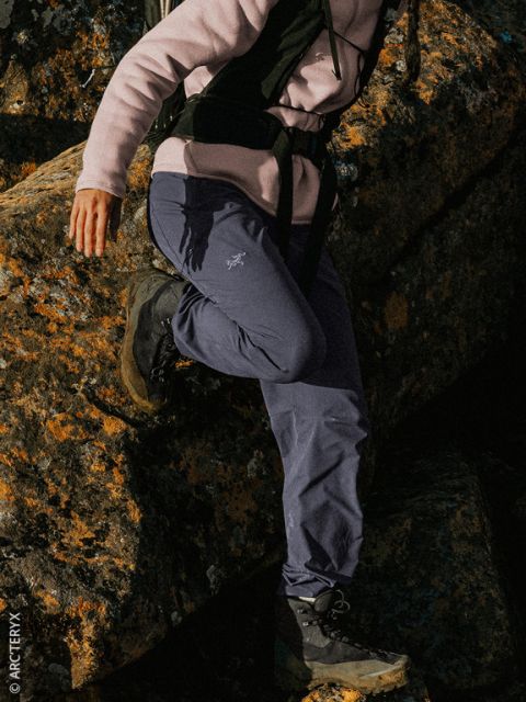 Eine Person in Outdoor-Kleidung klettert über mit Flechten bewachsene Felsen; sie trägt Wanderstiefel, eine blaue Hose und eine rosa Jacke.