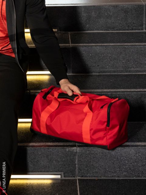 Eine Person in hellbeiger Jogginghose, schwarzen Turnschuhen und schwarzem Top hält einen glänzenden schwarzen Seesack. Sie stehen auf dunklen Holzdielen neben einem alten Scheinwerfer und gestapelten Koffern.