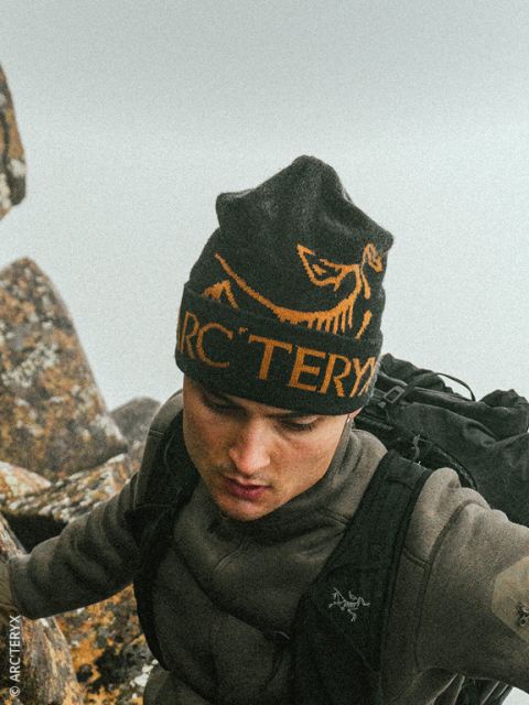 Eine Person in Outdoor-Kleidung und einer schwarzen Arc’teryx-Mütze klettert an einem bewölkten Tag über felsiges Gelände.