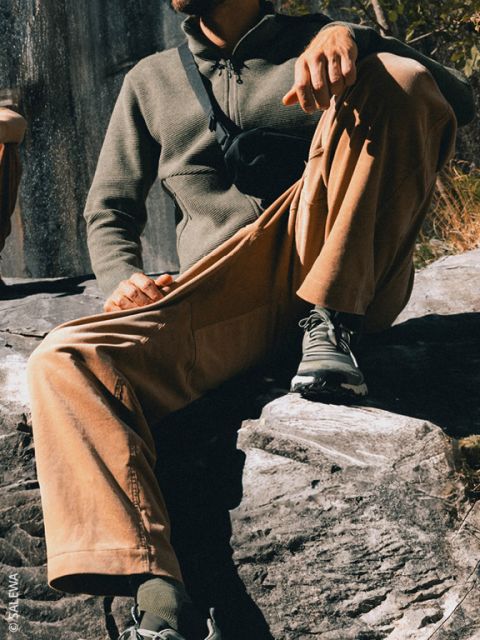 Eine Person sitzt draußen auf Felsen und trägt eine hellbraune Hose, ein grünes Oberteil, Turnschuhe und eine schwarze Umhängetasche; das Sonnenlicht wirft Schatten.