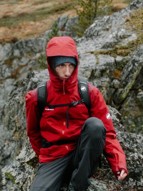 Eine Person in einer roten Kapuzenjacke und mit einem Rucksack sitzt draußen auf Felsen, im Hintergrund sind Bäume und felsiges Gelände zu sehen.