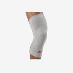 Eine Person, die eine graue Kniebandage mit einem kleinen roten Logo trägt, dargestellt mit angewinkeltem Knie vor einem schlichten weißen Hintergrund.