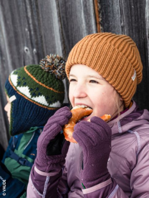 Zwei Kinder in Winterkleidung sitzen an einer Holzwand. Das eine isst eine Brezel, trägt eine braune Mütze und lila Handschuhe; das andere trägt eine grüne Mütze.