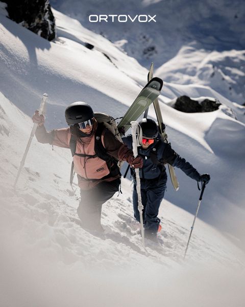 Drei Personen in Outdoor-Ausrüstung sitzen lächelnd und unterhalten sich auf felsigem Gelände in einer Bergregion. Die Sonne versinkt hinter den Gipfeln. Ein Rucksack und eine Thermoskanne stehen in der Nähe. Das Logo von Ortovox befindet sich oben rechts.