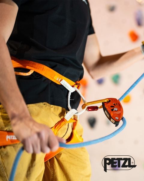 Person, die mit einem Klettergurt und einem Seil sichert und dabei ein orangefarbenes Sicherungsgerät verwendet; das PETZL-Logo ist in der rechten unteren Ecke zu sehen.