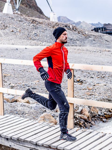 Ein Mann in einer roten Jacke, einer schwarzen Hose, Handschuhen und einer Mütze läuft auf einer Holzbrücke in einer felsigen, bergigen Gegend.