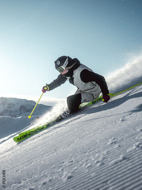 Ein Skifahrer in blauer Jacke und mit Helm fährt bei Sonnenuntergang einen verschneiten Berghang hinunter und wirbelt dabei Pulverschnee auf, während im Hintergrund schroffe, sonnenbeschienene Berggipfel zu sehen sind.