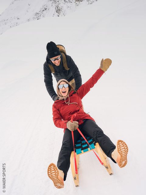 Zwei Personen in Winterkleidung ziehen Holzschlitten einen verschneiten Hügel hinauf, mit Bäumen und einem sonnenbeschienenen Berggipfel im Hintergrund. In der Ferne sind weitere Personen zu sehen.