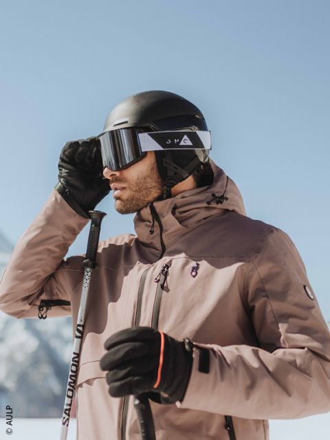 Nahaufnahme einer Person, die einen grauen Skihelm, eine reflektierende Skibrille und eine dunkle Jacke trägt, mit einer verschwommenen Schneelandschaft im Hintergrund.