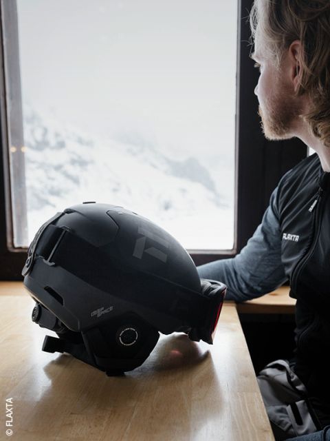 Eine Person mit hellem Haar und Bart sitzt an einem Holztisch am Fenster und blickt nach draußen auf verschneite Berge. Ein schwarzer Skihelm liegt vor ihnen auf dem Tisch.