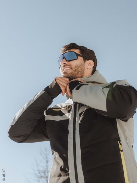 Ein Skifahrer, der einen weißen Helm, eine reflektierende Brille, eine schwarz-braune Jacke und Handschuhe trägt, schließt seine Jacke im Freien mit verschneiten Bergen im Hintergrund ab.