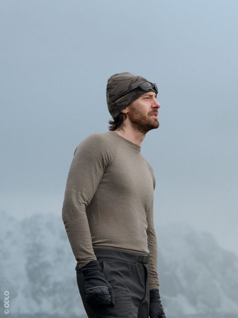 Ein Mann und eine Frau in legerer Winterkleidung sitzen im Freien an einer Steinmauer, lächeln und unterhalten sich. Ein Paar Skier lehnt an der Wand, und im Hintergrund sind schneebedeckte Berge zu sehen.