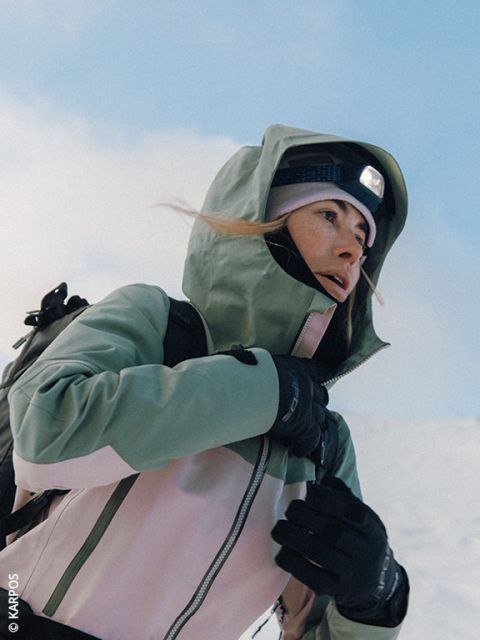 Eine Frau, die eine bauschige Dynafit-Jacke, eine Sonnenbrille und Handschuhe trägt, bereitet sich auf eine Wanderung in einer verschneiten Berglandschaft vor, mit schneebedeckten Gipfeln im Hintergrund. Sie trägt einen Rucksack und Skistöcke.