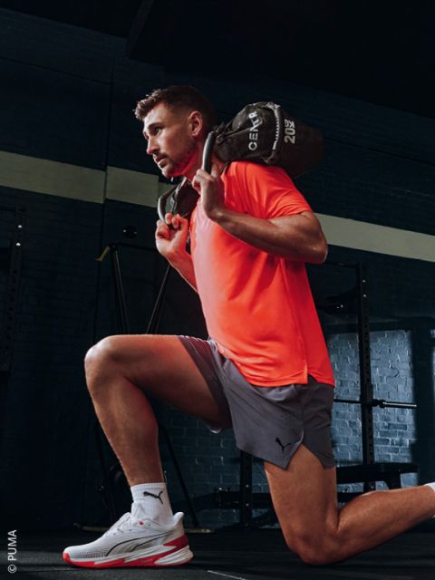 Ein Mann in einem orangefarbenen T-Shirt und grauen Shorts macht in einem Fitnessstudio einen Ausfallschritt, während er einen mit Gewichten gefüllten Sack über den Schultern hält.